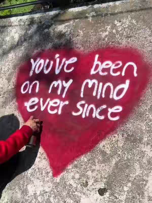 A close-up shot of a concrete wall with a heart-shaped graffiti. Inside the red-painted heart, the text "you've been on my mind ever since" is written in white, cursive letters. To the left, a person's arm with a red sleeve is visible, holding a spray paint can.