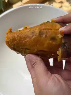 A hand is holding a cooked sweet potato in front of a white bowl. The sweet potato is cooked and has a vibrant orange color, with its skin partially removed. The background is slightly blurred but appears to be a kitchen setting.