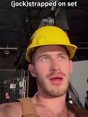 A man in a yellow hard hat and work attire looks up with an open mouth in a setting suggesting a film set. Text overlayed onto the image says '(jock)strapped on set'. The background is dimly lit with equipment visible.