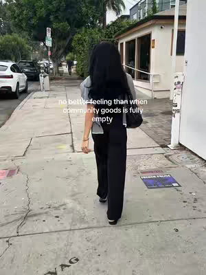 A person with long black hair walks away from the camera on a sidewalk. The person wears a gray top and black pants. Buildings and parked cars are behind the figure.