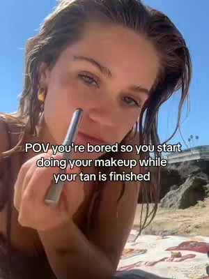 A selfie-style shot with a young woman at a beach. She holds a makeup applicator to her nose and looks at the camera. The background shows a bright blue sky and part of a sandy beach and some construction.