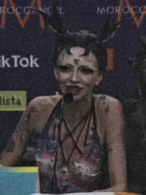A person with elaborate face makeup and hair accessories is positioned against a dark background. The image has a grainy texture. The individual is wearing a halter top. There are text overlay elements on the image.