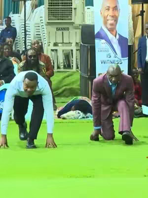 Two men are kneeling on a bright green surface with their hands placed on the ground, as if they're in a starting position for a race. A large banner with a man's face is in the background, and other individuals are also present.