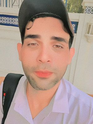 A man with dark hair and a beard is wearing a black baseball cap and a white collared shirt. He has a black backpack. The background has a white building and a blue decorative pattern. The lighting is bright.