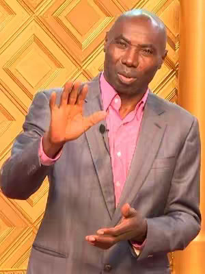 A man with dark skin, wearing a gray suit over a pink shirt, is positioned in front of a patterned golden backdrop, with one hand raised as if speaking or emphasizing a point.