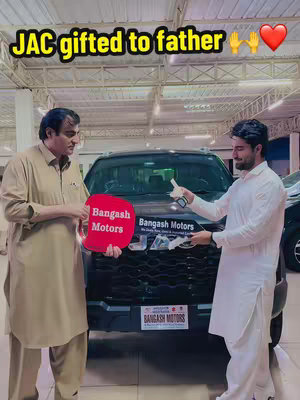 A man in a light brown shirt and pants is holding a red sign that says "Bangash Motors" in front of a black SUV, giving it to another man in a white shirt. The image is indoors with bright lighting, and there are other cars and windows in the background. The man on the right is holding a key.