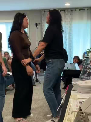 Two women are in a living room setting. One in a black shirt and jeans is touching the arm of another wearing a dark long-sleeve top and black pants. Other people are visible in the background.