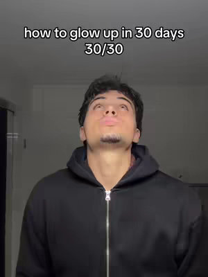 A young man in a black hoodie looks upwards with his lips pursed as if he's about to blow a kiss. Above him, in a simple font, text reads, "how to glow up in 30 days 30/30". The background appears to be a plain, light-colored wall, and the lighting is diffused.