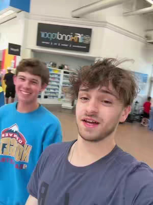 Two young men inside of what looks like a retail store, one is filming with one hand and has an open mouth talking into the camera, the other guy on the other side smiling at the camera. the store has white ceiling, a sign with writing and another sign on the left.