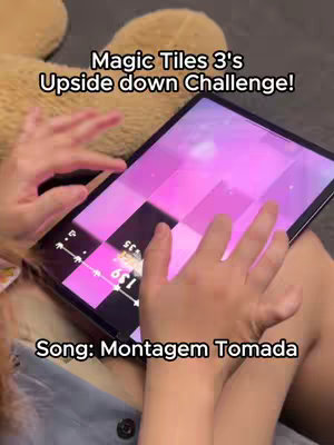 The cover image shows a person holding a tablet that displays a mobile game. The person's hands are holding the tablet, and the game interface with black and pink tiles is on the screen. The background is a blurry image of a teddy bear and possibly the person's legs. Text overlays near the top indicate the name of the game and a challenge related to it and text overlays near the bottom indicate a song name.