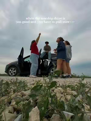 A group of young people are gathered near a black car parked on a gravel road with a cloudy sky overhead. The backdrop includes fields and a distant treeline. One person is standing in the car with their arm raised, as if reaching upwards.