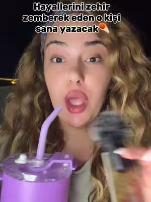 A woman with long curly hair is holding a purple drink container and a microphone, her mouth open as if she is speaking. Text is overlaid at the top of frame.