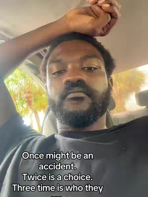 An individual with a beard and dark hair, wearing a black t-shirt, is seated inside of a vehicle. The person's right arm is placed above their head, next to the vehicle's ceiling. Text overlays the lower center of the image.