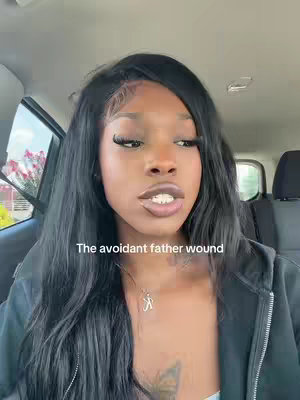 A woman with long black hair is sitting inside a car. She's wearing a black hoodie and a necklace with a pendant. Text overlays on the image says "The avoidant father wound". The car interior, including a headrest, is visible in the background