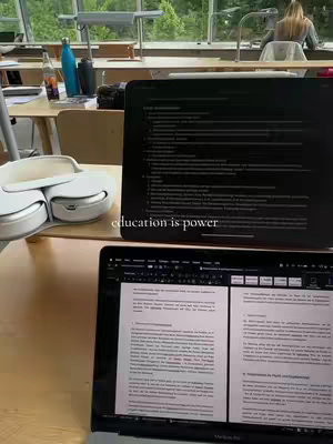 An office setting with a close up shot of a laptop and iPad on a wooden desk. A pair of white headphones sits on the desk alongside the iPad. In the background, are high windows exposing an outdoor natural landscape and a blurred figure of a person.