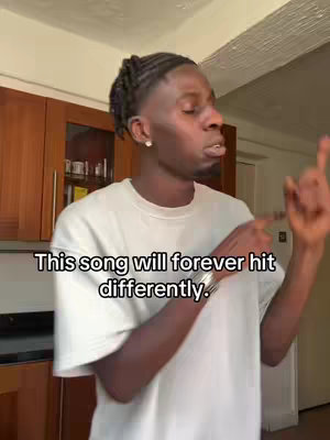 A man with braided hair is in a kitchen setting. Text overlayed on the video says 'This song will forever hit differently'. He raises his right hand with the index finger pointing up and is looking off to the right.