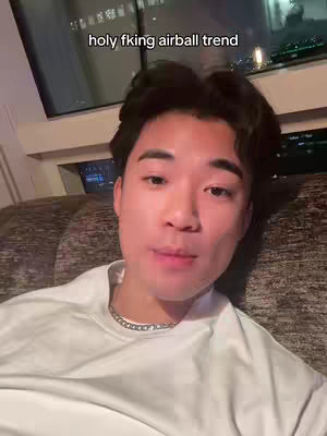 A young Asian man looks directly at the camera. The background is a dark room with a window, where lights can be seen.