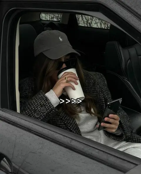 A woman sits inside a car, holding a white coffee cup and a phone and looking at the phone. She wears a gray cap, sunglasses, a dark patterned coat, and a gray shirt. The car interior is dark, a few trees are visible blurred in the background, and there is text overlay in the image.