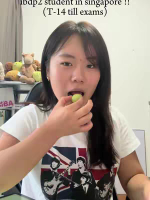 A young woman is shown in a head-and-shoulders shot, holding a green grape and about to put it in her mouth. She wears a white t-shirt with a graphic featuring The Beatles. Above her head in the image, white text reads, 'ibdp2 student in singapore !! (T-14 till exams)'. The background is a room interior with a shelf holding plush toys and a partially visible computer screen.