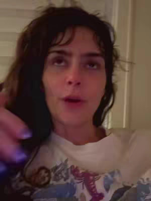 A woman with dark, wavy hair is looking up towards the top right of the frame, with her mouth slightly open as if she is speaking. She is wearing a white t-shirt with a colorful design. The background is blurry, indicating an indoor setting.