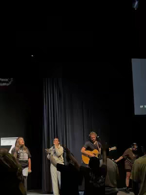 The cover image shows a stage with several people performing in front of an audience. In the foreground, people raise their hands in worship. In the center, a person plays a guitar. To the left, there is a woman singing into a microphone. Behind them is a dark curtain, behind which are lights. A screen on the stage displays text.
