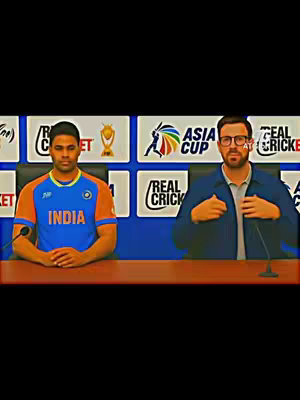 A vertical video cover featuring two men seated at a table. The man on the left is wearing an Indian cricket team jersey. The man on the right is wearing a navy blazer and glasses. Behind them are promotional boards with the "Asia Cup" and "Real Cricket" logos.