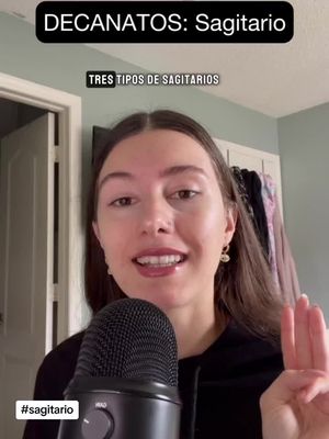 A woman in a black hoodie is positioned in front of a neutral-toned background, likely indoors. Above, a banner reading 'DECANATOS: Sagitario' is present. The hook of the video shows 'TRES TIPOS DE SAGITARIOS'. The woman is holding an open microphone, while the woman gestures three fingers up. The video appears to discuss topics related to astrology.
