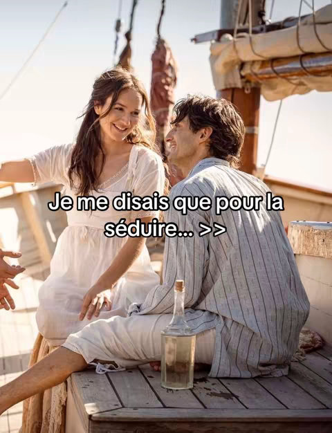 A couple is seated on a wooden deck of a boat, smiling at each other. The woman wears a white dress, and the man is in a striped shirt and white pants. A bottle sits on the deck. The background shows a sunny sky and the boat's rigging.