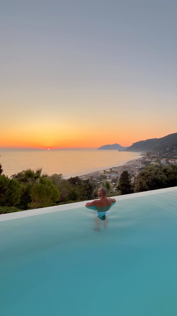 A tranquil scene featuring a man in a pool overlooking a beach at sunset. The man is seen from the back, with his arms resting on the pool's edge, facing towards a vibrant sunset over the sea. The water in the pool is a clear, turquoise color, contrasting with the orange and yellow hues of the setting sun. The background shows a coastline with buildings and lush greenery. The image evokes a sense of relaxation and luxury.