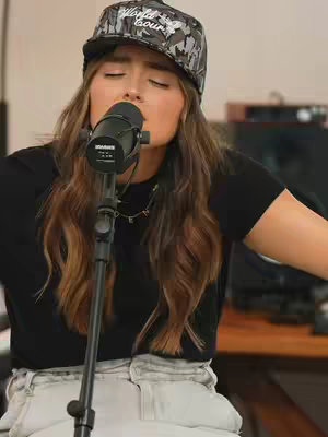 The image is a medium shot featuring a young woman with long, wavy brown hair. She wears a camouflage-patterned baseball cap with the words "World Court" on it and a black t-shirt. She's positioned behind a microphone, eyes closed, seemingly singing with emotion. The background is blurred, suggesting a studio setting. The lighting appears to be soft and focused on the presenter.