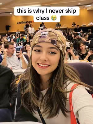 A young woman with long brown hair and a camo beanie smiles in a large lecture hall. A text overlay arches above her: "this is why I never skip class 😜". The hall is filled with other students, some holding phones. The image is well-lit, with a warm color palette.