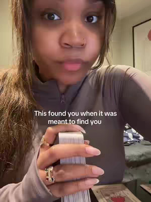 A woman with brown hair and a neutral expression is holding a deck of tarot cards. She has multiple rings on her fingers. Text overlayed on the image says, “This found you when it was meant to find you.”