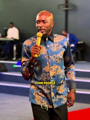 A man with a bald head is speaking into a microphone while on stage. He is wearing a blue patterned shirt over black pants. Beneath him are red steps, and the background is blurred, suggesting a church or auditorium setting.