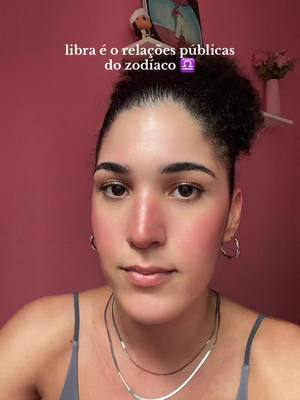 A selfie featuring a woman with dark curly hair pulled back. She has a neutral expression, wearing a gray top and layered necklaces. The background is a solid pink wall with a small shelf holding flowers. Text in the image reads: libra é o relações públicas do zodíaco ♎. The overall tone is related to astrology.