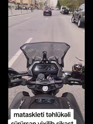 A first-person view from a motorcycle, driving down a city street. The foreground shows the motorcycle's handlebars, dashboard, and windshield with a blurred background of cars and buildings. The sky is overcast.