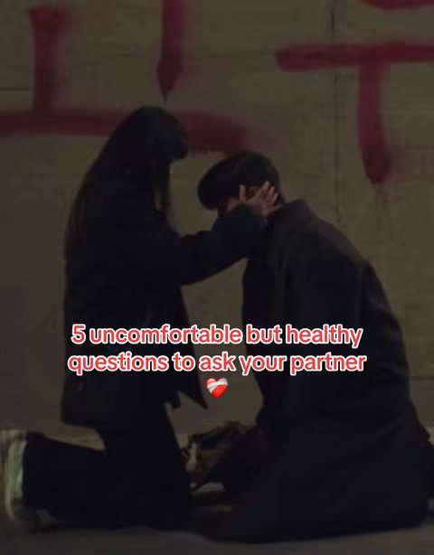 The image features two people, likely a couple, in an intimate setting. A woman is kneeling, her hand on the man's face, with both appearing to be deep in conversation. The setting is a dimly lit, urban-looking space, with the text overlay stating '5 uncomfortable but healthy questions to ask your partner.'