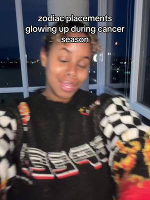 A person with light skin and short, styled hair is centered in the frame, wearing a dark sweater with a decorative design. The background shows a nighttime cityscape through a large window. The text overlay reads 'zodiac placements glowing up during cancer season'.