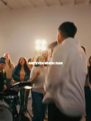 The image shows a group of people in a brightly lit room, seemingly singing or chanting.  In the foreground, a person with short hair, wearing a white top and moving. The backdrop is plain and the overall lighting is warm.