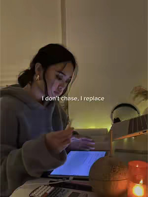 A young woman sits at a desk, illuminated by warm lighting, possibly studying. She has dark hair pulled back, wearing a gray hoodie, and is looking down at a tablet. Text overlayed reads: 'I don't chase, I replace'. A lamp is on the desk, and a candle burns in the corner.
