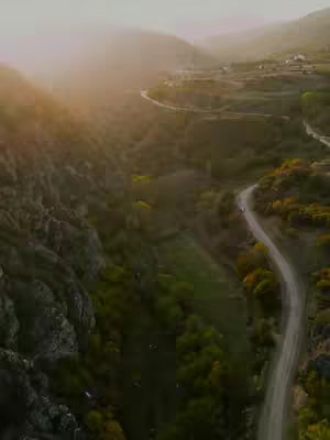An aerial view of a winding road through a valley. The valley is filled with green foliage and rolling hills. The sky is a hazy yellow.