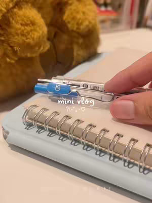 A hand holds a pen over a spiral notebook. The background includes a fuzzy stuffed animal and hints of other objects. The notebook is light blue, and the pen has blue accents. The text "mini vlog" is overlaid on the image in a playful font.