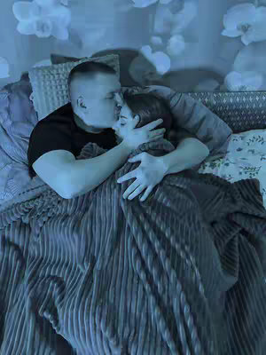 A man kisses a woman on the forehead while they are lying in bed together. They are covered by a blanket, and the scene is dimly lit with a blue-toned hue.