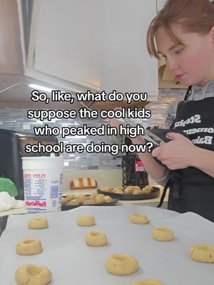 A woman is in a kitchen, likely baking. She is standing in front of a counter with a baking sheet of cookies. Text overlay reads "So, like, what do you suppose the cool kids who peaked in high school are doing now?"