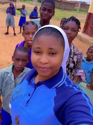 A selfie taken in an outdoor setting, featuring a woman in the foreground surrounded by several children. The woman is central, with a light skin tone, wearing a blue shirt with a white head covering. The children are of various ages and ethnicities, some with school uniforms. They all are looking towards the camera. The background shows a building, a sandy ground.