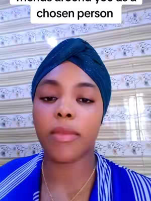A woman with a dark blue head wrap poses for a selfie. The shot is a close-up, highlighting her face. She is wearing a blue and white striped top with a gold necklace. The background is a patterned wall.