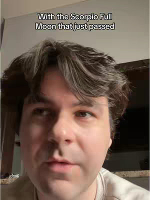 A man with short, wavy, dark hair looks into the camera with a neutral to slightly questioning expression. Added text overlay above reads, "With the Scorpio Full Moon that just passed". The background is dim and out of focus.
