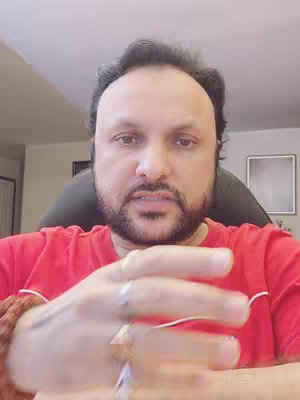 A middle-aged man with a beard and short dark hair is centered in the frame, wearing a red shirt. He looks directly at the camera with a slightly quizzical expression, with his hand raised.