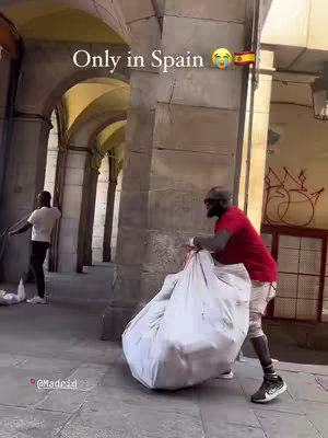 A man in street clothes, in a red shirt and shorts, is hastily gathering items into a large white bag with a partner standing in the background. They are in a stone-paved outdoor space. Text appears to be added to the image in the top left corner. A blurred view of an arched structure forms the backdrop.