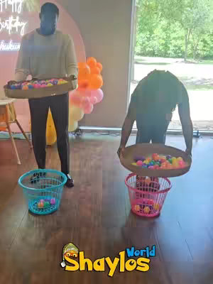 The image shows two people playing a party game. One person is standing holding a tray with colorful balls, while the other is bent over a basket. Baskets and balloons are in frame.