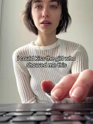 A woman with dark hair and silver hoop earrings looks directly at the camera, positioned in front of a light-colored wall. The text overlay reads, 'i could kiss the girl who showed me this'. Her hand is visible, touching a laptop keyboard in the foreground creating a sense of interaction and engagement.